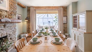 A dining room with a long table set for six, stone walls, a large window, and a view of the sea.
