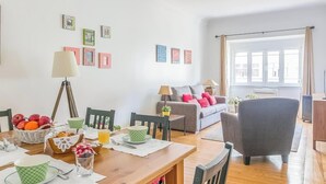 A cozy living room with a dining table, a sofa, and a chair. There are framed pictures on the wall, a lamp, and a window with curtains. - Casa da Avenida III Lisboa (Lisbon)
