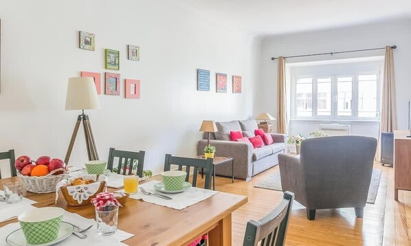 A cozy living room with a dining table, a sofa, and a chair. There are framed pictures on the wall, a lamp, and a window with curtains.