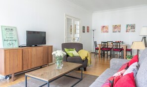 Una sala de estar con un sofá, una mesa de centro, un televisor sobre un mueble de madera, un área de comedor con sillas y una mesa, y obras de arte en la pared.
