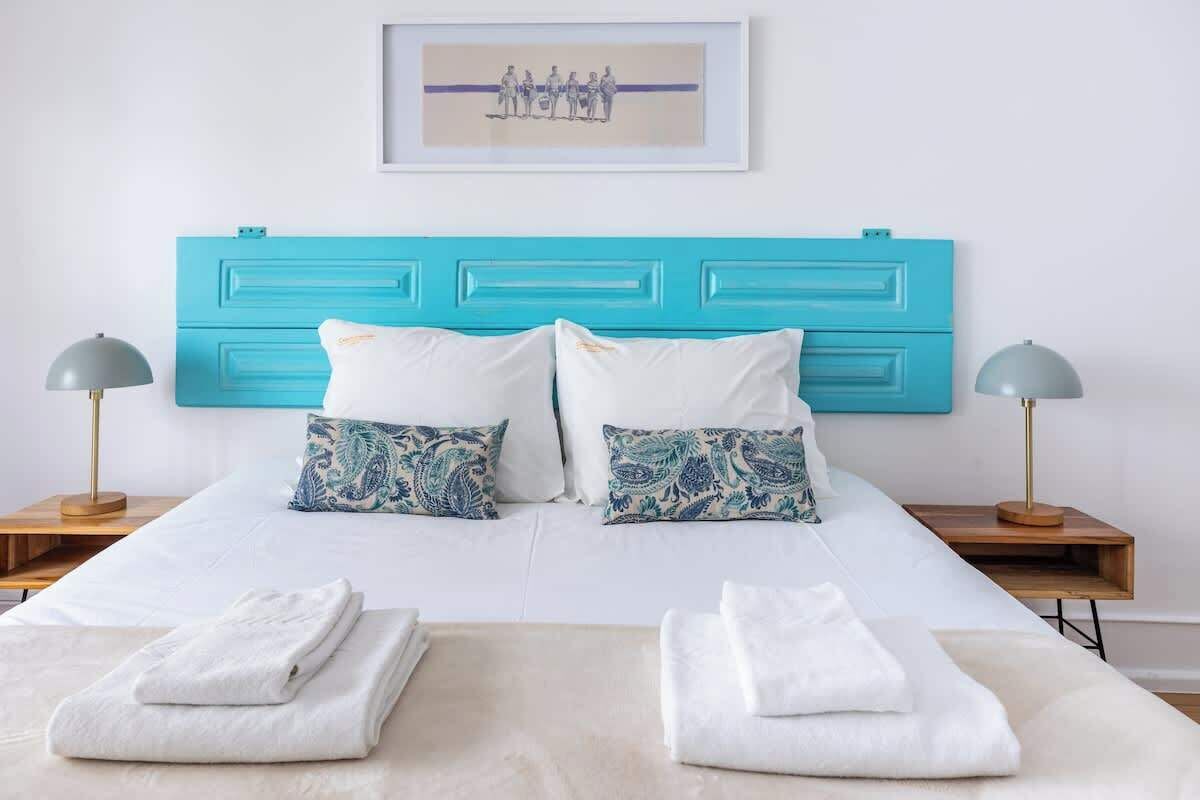 A bed with a turquoise headboard, white bedding, two patterned pillows, and two bedside lamps.