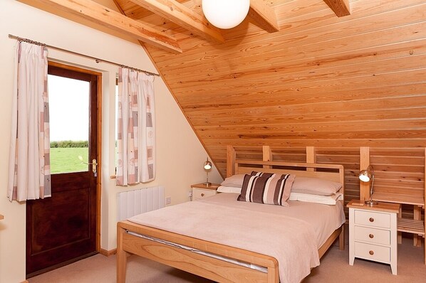 Un dormitorio con techo inclinado, cabecera de madera, cama con colcha color crema, dos mesitas de noche con lámparas y una ventana con cortinas.