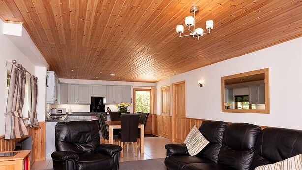 A modern living room with a wooden ceiling, black leather sofas, a dining table, and a kitchen in the background.