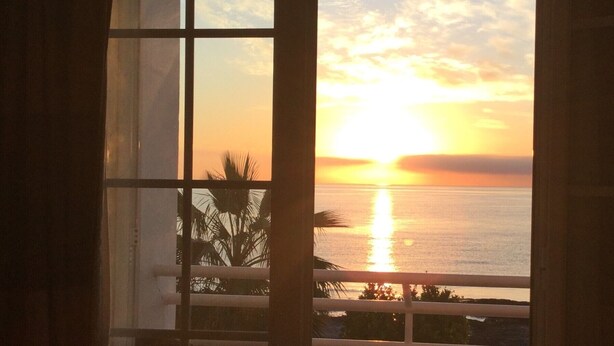 A balcony with a view of the sunset over the ocean, a table, and two chairs.