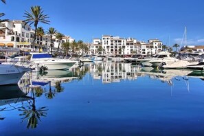 A marina with boats docked, surrounded by multi-story buildings with balconies, palm trees, and clear blue skies. - 2 Bed, 2 Bath Apartment with a huge balcony several seating areas  (La Duquesa)