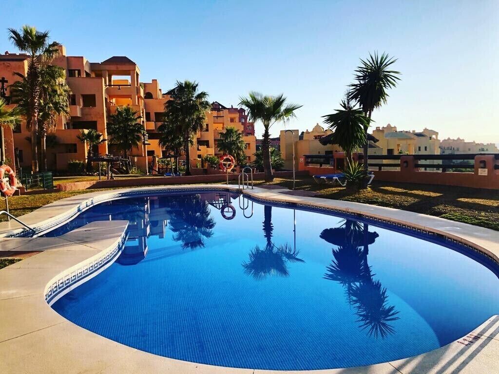 A swimming pool with a curved edge, surrounded by palm trees and a building with multiple balconies in the background.