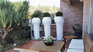 Een terras met een houten tafel, wit beklede zitplaatsen en drie grote witte plantenbakken met planten.