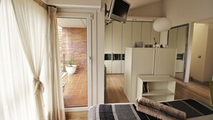 Een moderne slaapkamer met een bed, een aan de muur bevestigde televisie, een badkamer met spiegel en uitzicht op een bakstenen muur en een potplant buiten.