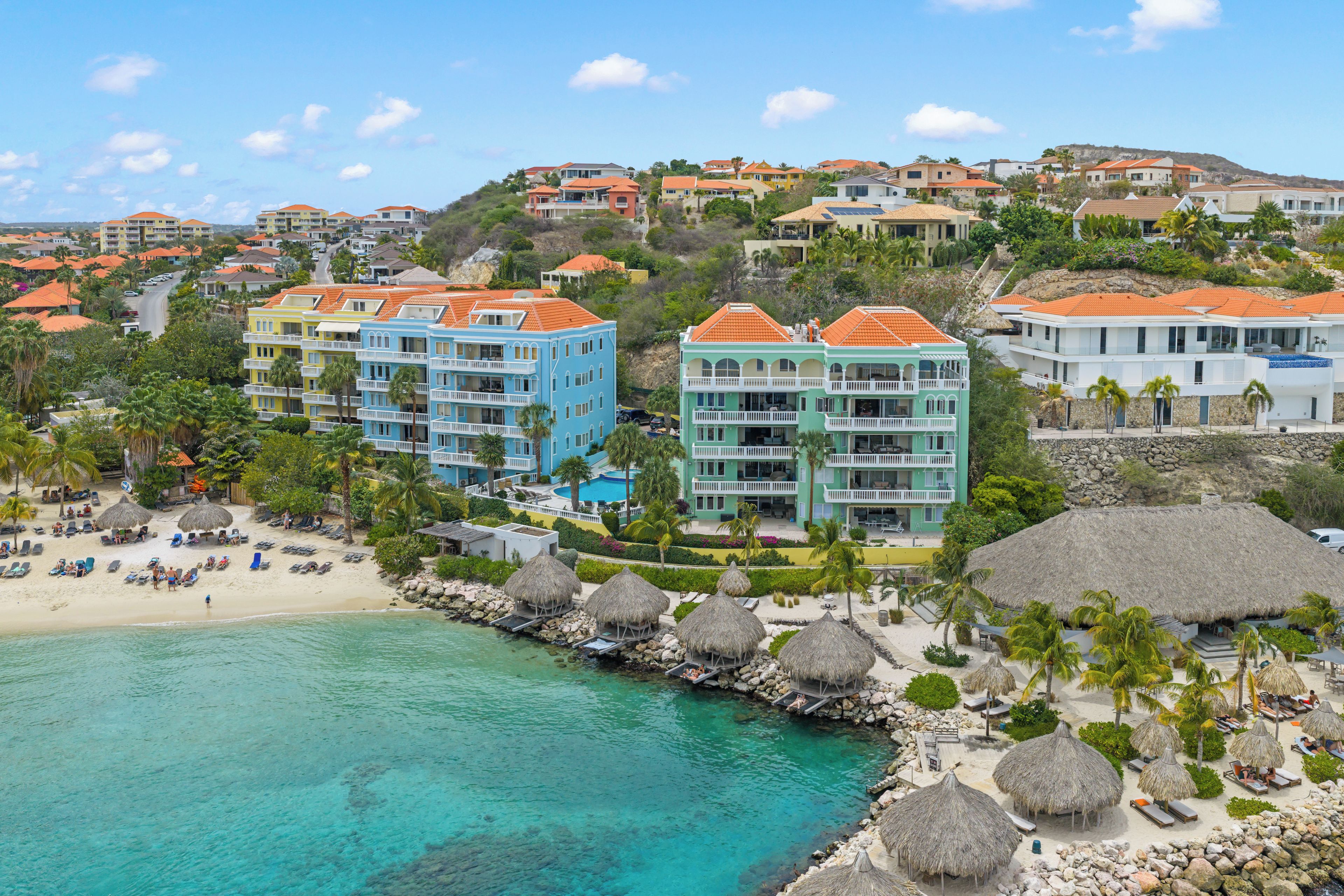 A coastal resort with multiple-story buildings, a beach with thatched umbrellas, and clear turquoise waters.