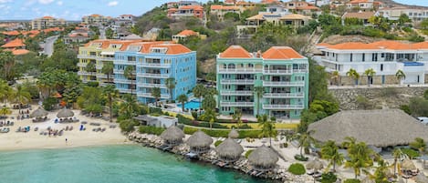 A coastal resort with multiple-story buildings, a beach with thatched umbrellas, and clear turquoise waters.