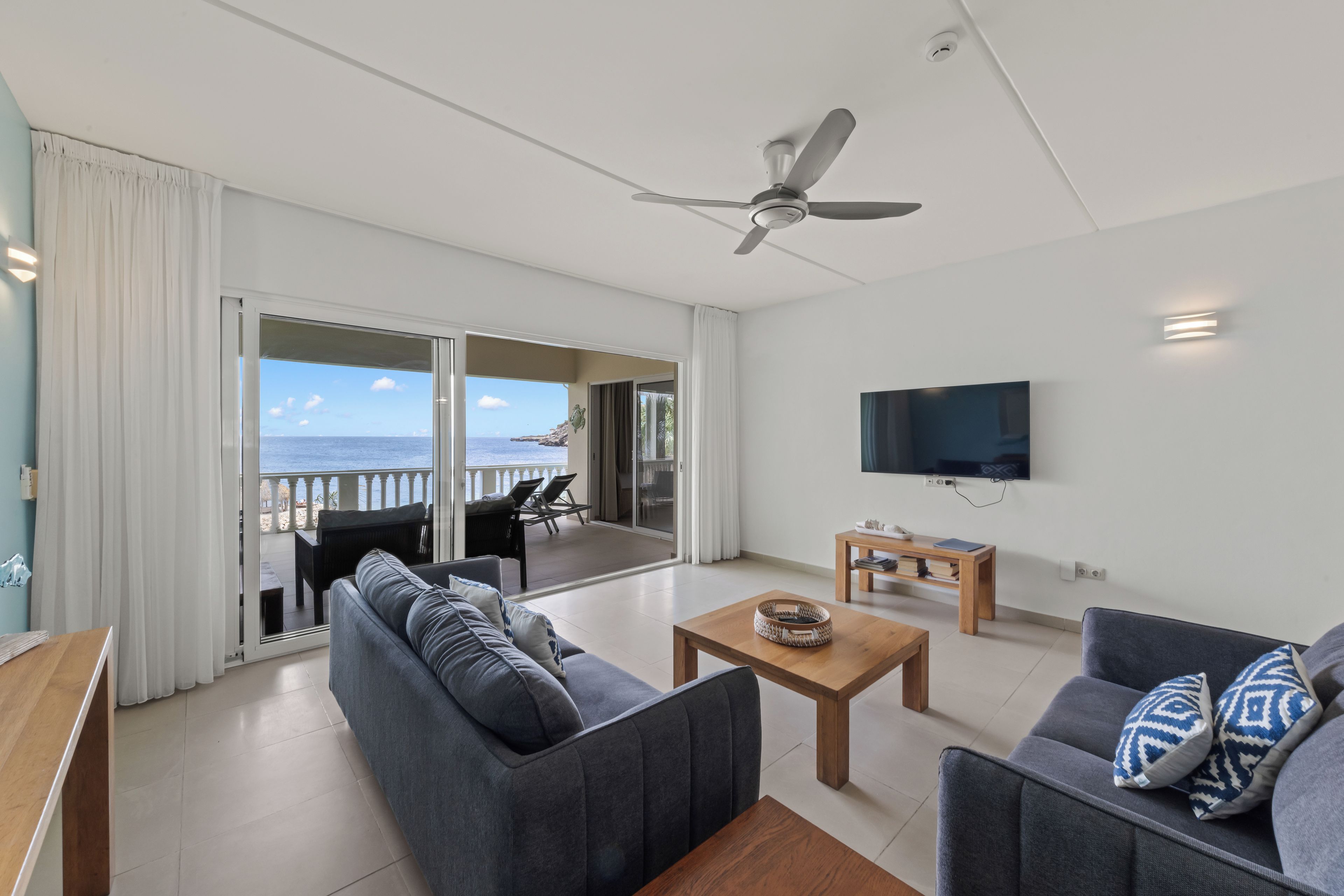 A modern living room with a sofa, a coffee table, and a TV. There is a balcony with a view of the sea.