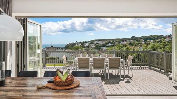 Une terrasse en bois avec des sièges d’extérieur, une table sur laquelle se trouve un panier de fruits, et une vue sur une ville située sur une colline.