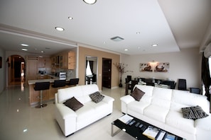 A modern living room with two white sofas, a coffee table, and a kitchen area in the background.
