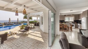 A modern kitchen with a dining area, a glass sliding door leading to a balcony with a pool, and outdoor seating. - Situated in a quiet development, this front line villa has views to die for (Protaras)