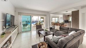 A spacious living area with a large sofa, a coffee table, and a TV on the wall. There is a sliding glass door leading to a balcony with a pool view. - Situated in a quiet development, this front line villa has views to die for (Protaras)