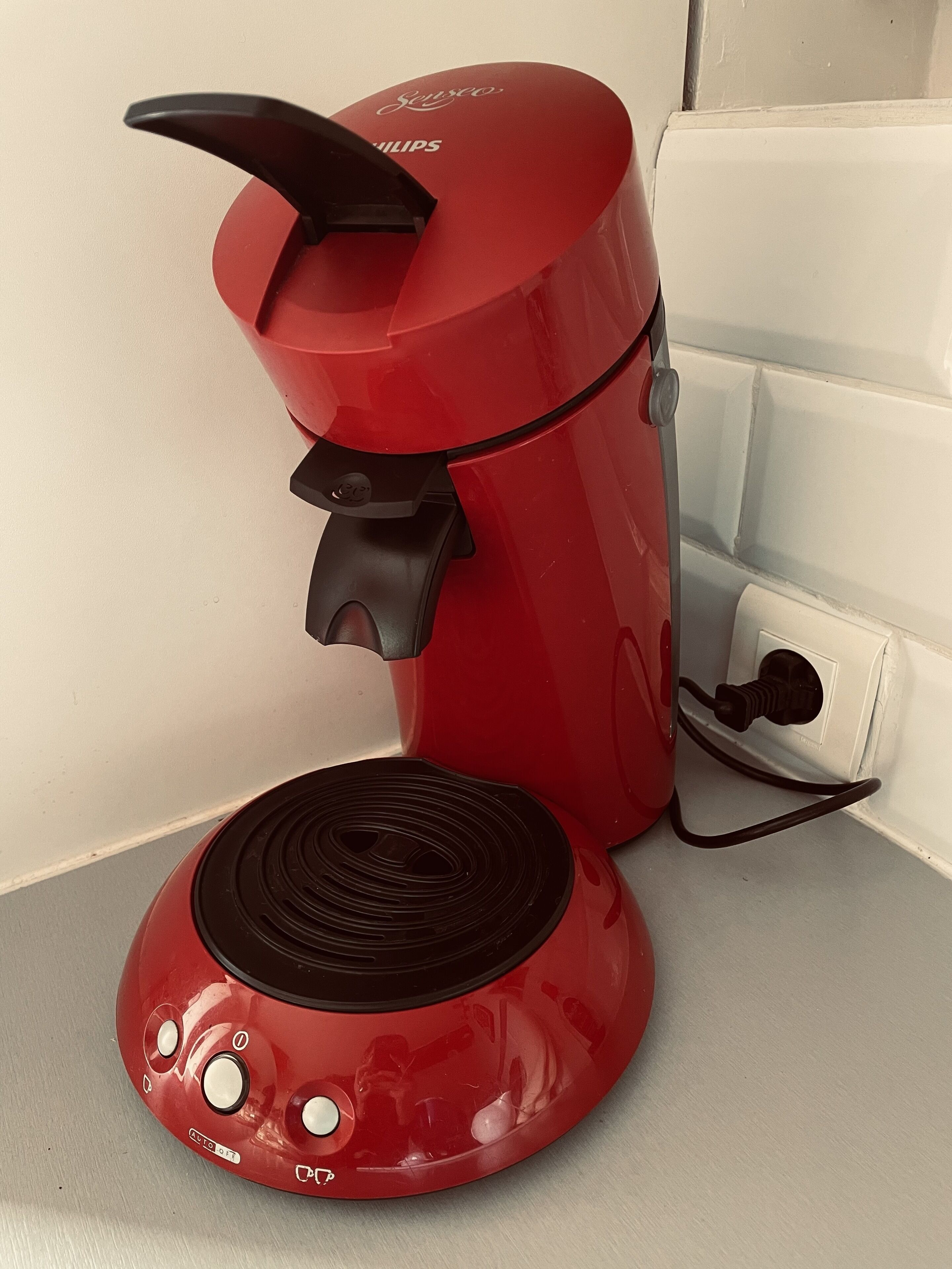 A red coffee maker with a black base and a power cord plugged into a wall socket.