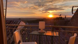 A balcony with two white chairs, a table, and a lamp. The view includes a sunset over a cityscape with buildings and a distant horizon.