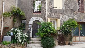 A rustic stone building with a black iron gate, a small arched window, and a door with a yellow shutter. There are potted plants and flowers around the entrance.