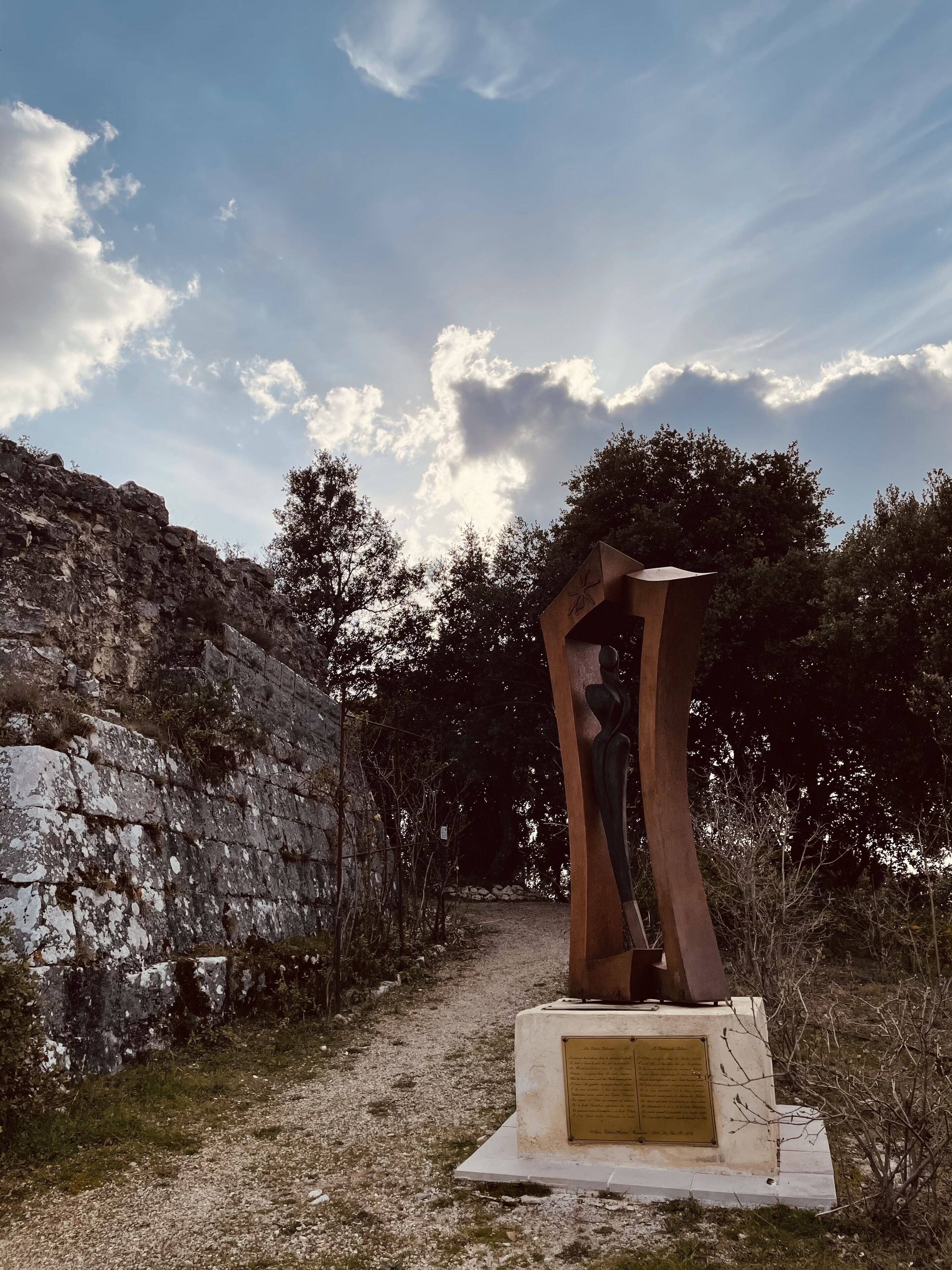 Eine Skulptur in einer natürlichen Umgebung mit einer Steinmauer und Bäumen im Hintergrund.