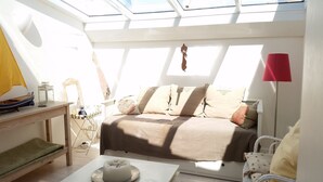 Un dormitorio iluminado por el sol con cama, una mesita, una silla y un tragaluz.