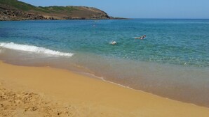 Une plage avec une eau d’un bleu limpide, un rivage sablonneux et un paysage vallonné en arrière-plan.