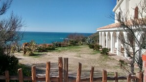 Une vue sur la côte, avec un sentier sablonneux menant à un bâtiment blanc orné de colonnes, entouré d’une végétation désertique.