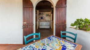 Un espace extérieur aménagé dans un style rustique, avec une table carrelée et deux chaises, qui donne accès à un intérieur chaleureux comprenant un canapé et des étagères.