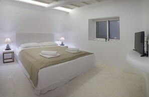 Ein Schlafzimmer mit einem großen Bett, zwei Nachttischen mit Lampen, einem Fenster mit Jalousien und einem an der Wand befestigten Fernseher.