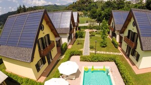 Un complexe hôtelier comprenant plusieurs maisons dotées de toits inclinés recouverts de panneaux solaires, d’une piscine et offrant une vue sur une colline lointaine surmontée d’une structure ressemblant à un château.