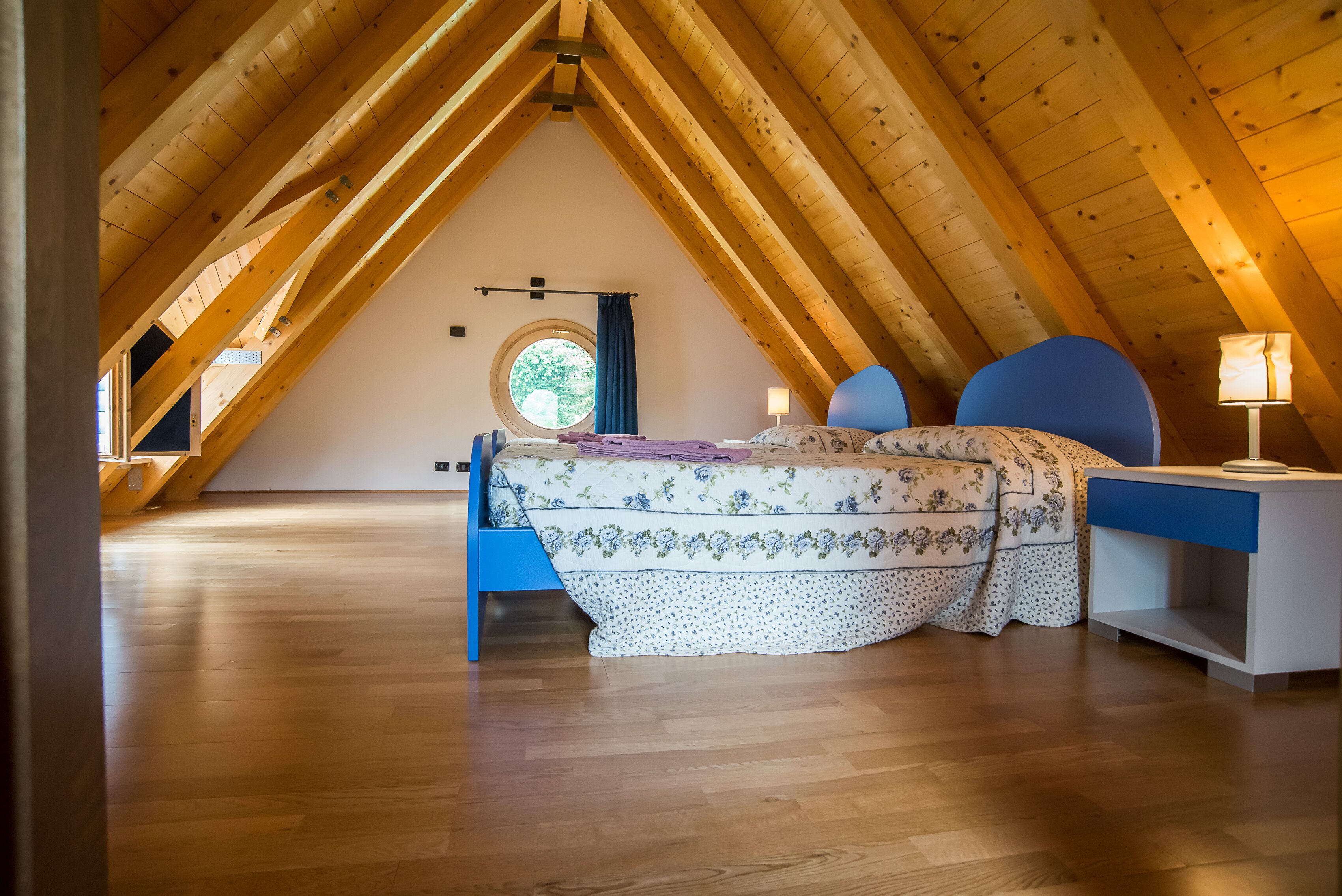 A bedroom with a wooden ceiling, a bed with floral bedding, a blue chair, and a nightstand with a lamp.