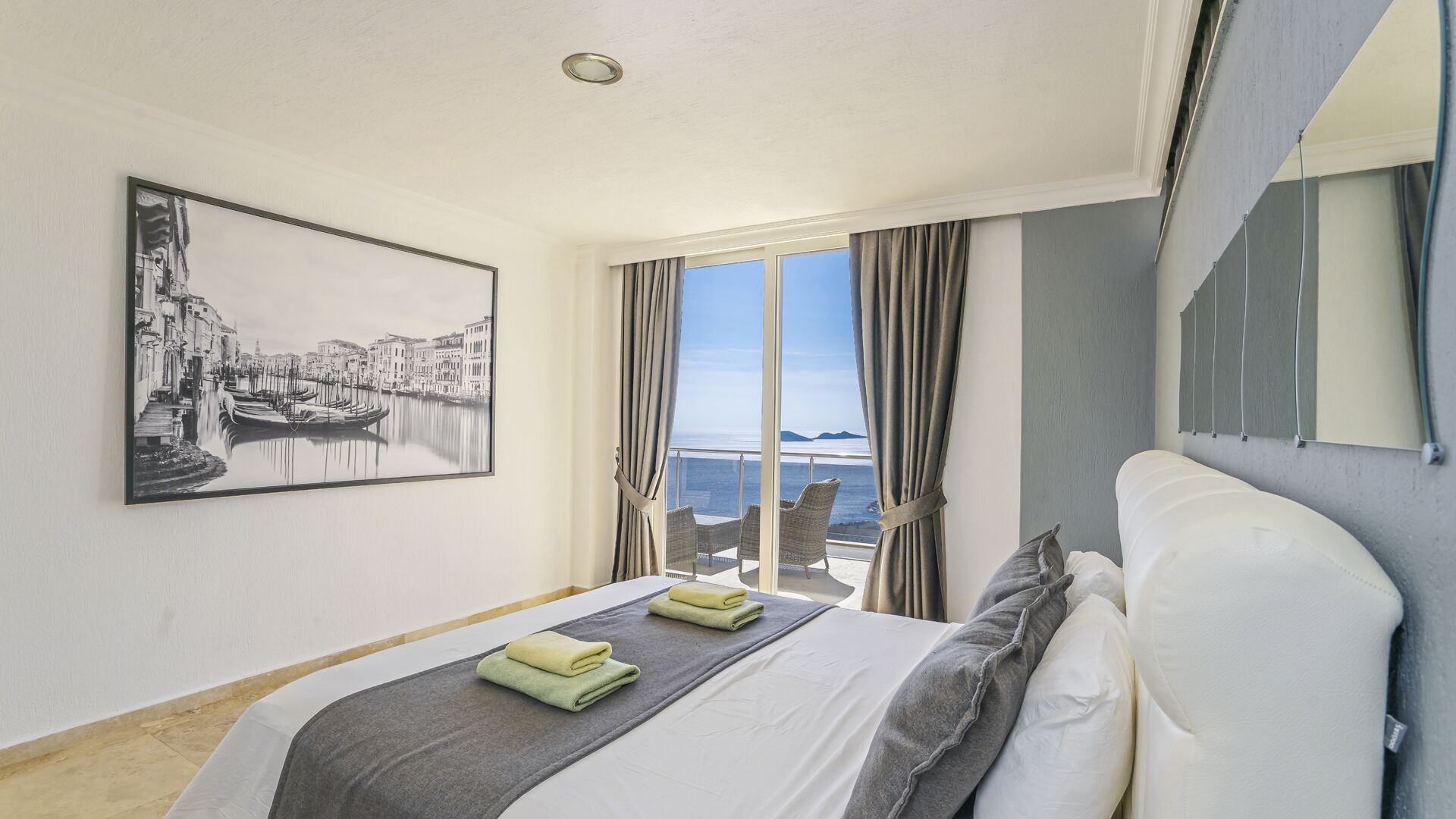 A hotel room with a bed, a large window with curtains, a view of the sea, and a framed picture on the wall.
