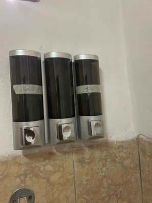 Kolme sylinterinmuotoista annostelijaa, joissa on merkinnät ”SHAMPOO” ja ”GEL”, on kiinnitetty seinään.