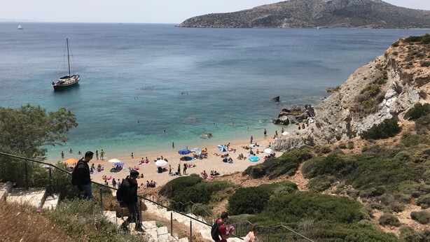 A coastal beach scene with clear waters, a sailboat, and people enjoying the beach. - Elea Athens Apartment (Athina)