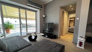 A modern living room with a sofa, a glass coffee table, a flat-screen TV, and a balcony with a potted plant. - Elea Athens Apartment (Athina)