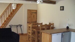 A compact living space with a wooden staircase, a dining area with wooden chairs, a small kitchen with a refrigerator, and a black sofa. - Fully Furnished 2 Bed Gite Situated In 3 Acres (Arné)
