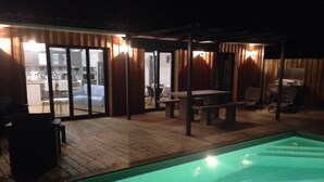 Une piscine avec une terrasse en bois, un patio couvert doté de tables et de chaises, et une vue sur la cuisine à travers des portes vitrées.