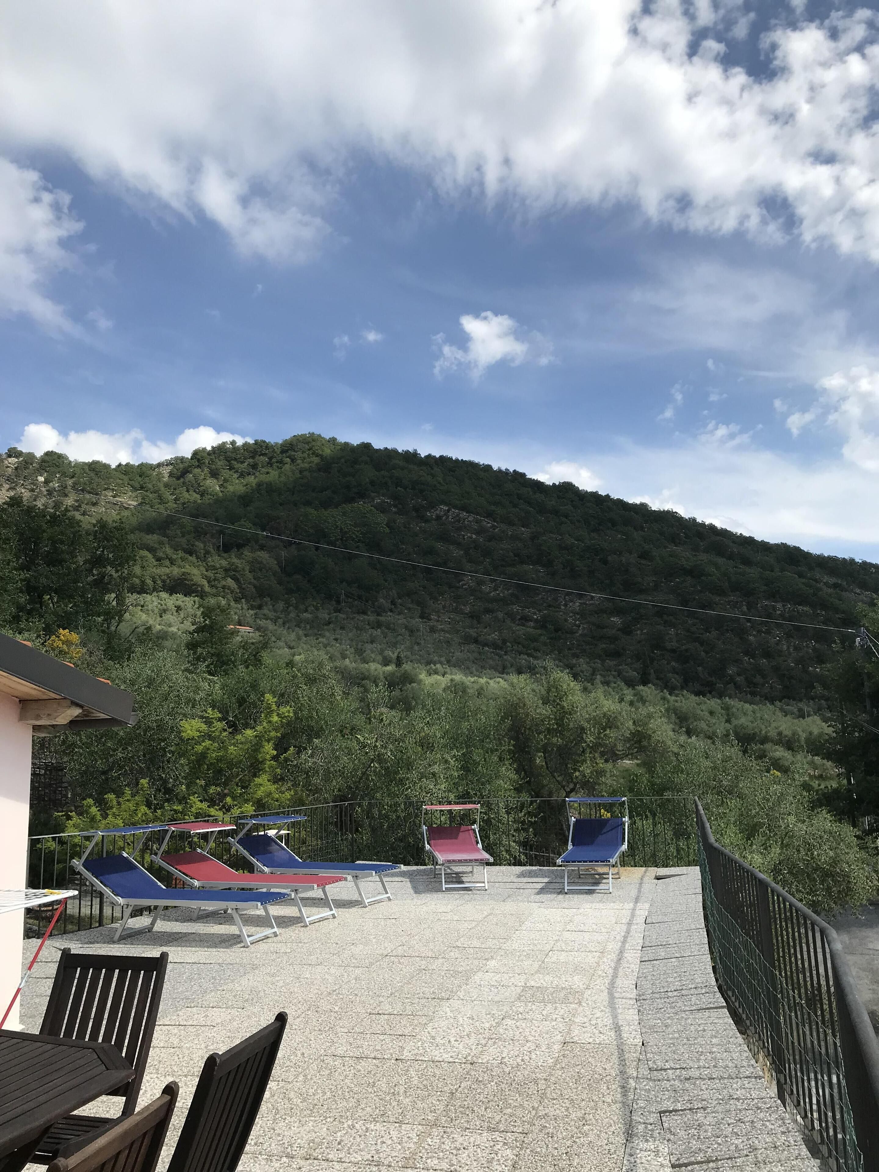 A terrace with sun loungers and a table, overlooking a forested hillside.