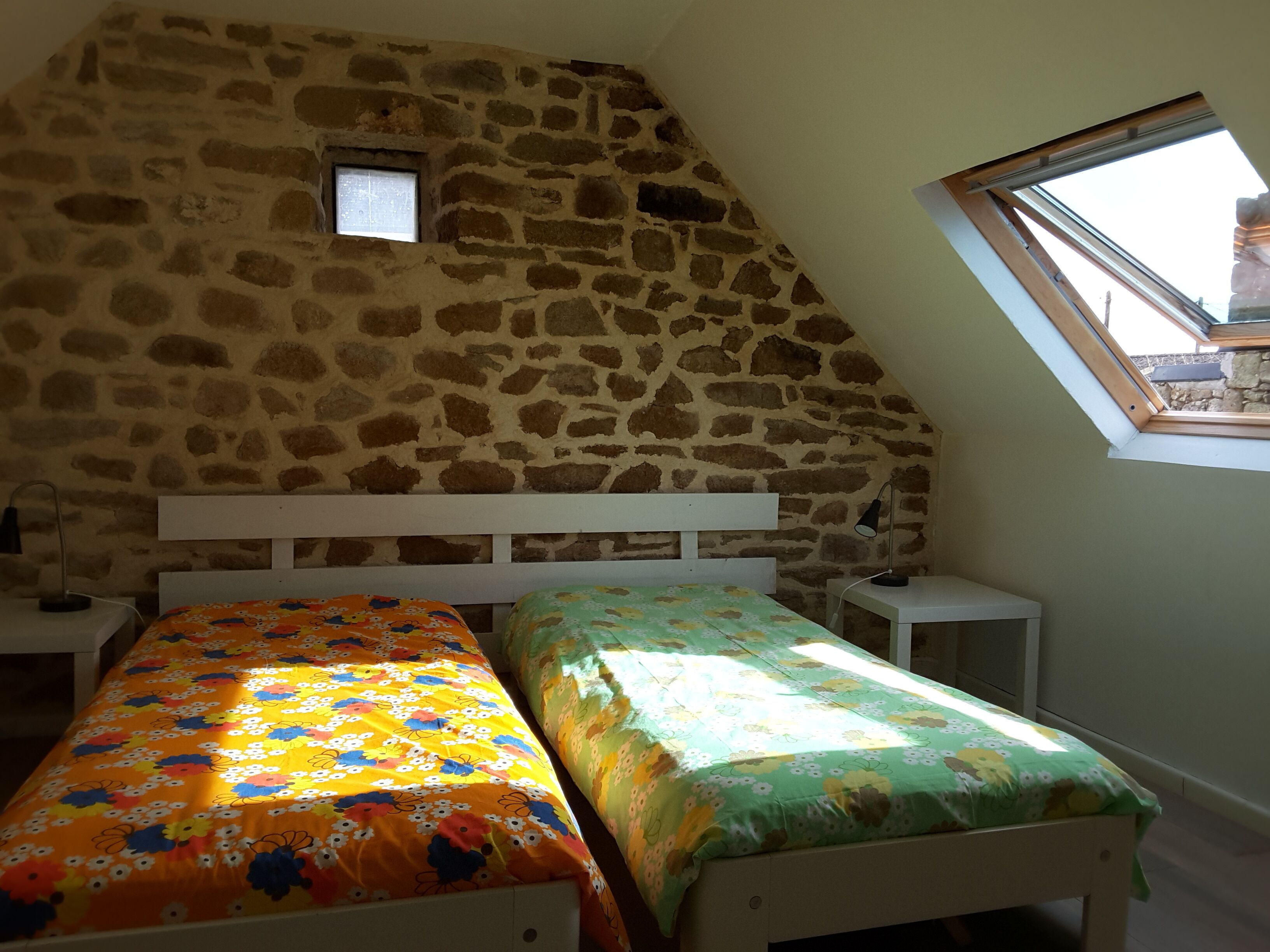 A room with a stone wall, a sloped ceiling, and a skylight. There are two beds with floral bedding.