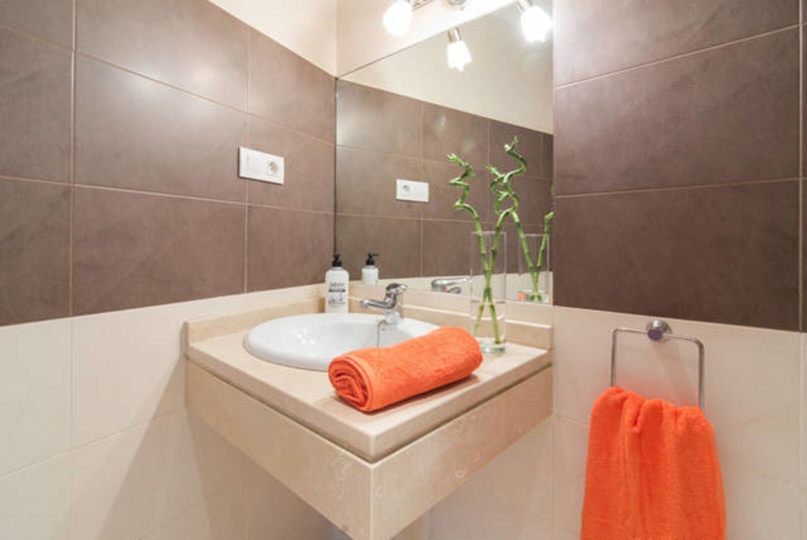 Un baño con lavamanos blanco, un espejo, un perchero con una toalla naranja y una planta pequeña.