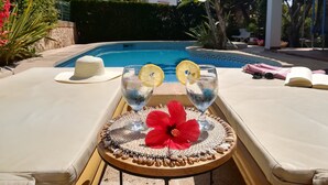 Escena junto a la piscina con dos vasos de agua, una flor roja y un sombrero blanco sobre una mesa.