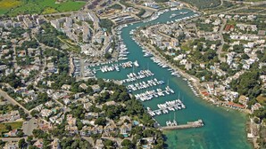 Vista aérea de un pueblo costero con un puerto deportivo, edificios residenciales y zonas verdes.