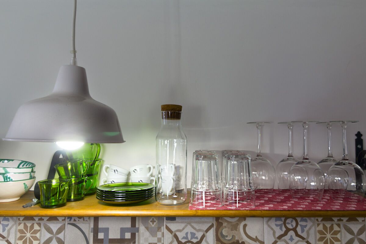 Uma bancada de cozinha com diversos copos de vidro, incluindo copos verdes e taças de vinho, e uma luminária pendente branca.