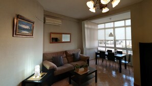 A living room with a sofa, coffee table, and dining area with chairs. There is a chandelier, a painting, and a window providing natural light.