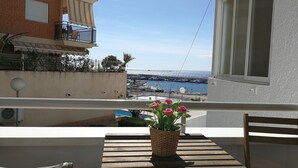 A balcony with a wooden table and chair, a potted plant, and a view of the sea and a bridge.