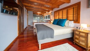 A bedroom with a wooden ceiling, a large bed with blue and white pillows, a bedside table with a lamp, and a view of the kitchen through a glass door.