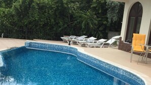 Une piscine dont le fond est recouvert de mosaïques bleues, entourée de chaises longues et bordée d’un bâtiment à la façade blanche.