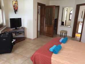 A hotel room with a bed, a TV, a desk with books, a suitcase, and a door leading to a bathroom.