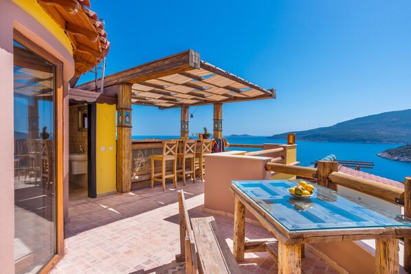 Een houten terras met een glazen tafel en stoelen, met uitzicht op een heldere blauwe zee.