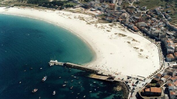 Vista aérea de un pueblo costero con playa de arena, un muelle y numerosos barcos.