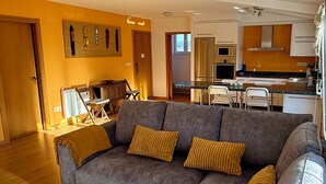 Una sala de estar moderna con un sofá gris, un reposapiés estampado y una cocina al fondo.